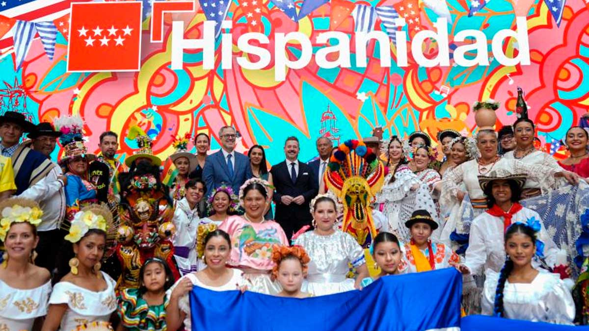 La imagen muestra a un gran grupo de personas con trajes típicos de diferentes países hispanos posando frente a un colorido mural con el texto "Hispanidad". El ambiente es festivo y alegre, reflejando la diversidad cultural y la importancia de la programación de la Hispanidad 2026. Al fondo se aprecian banderas y elementos decorativos alusivos a la celebración.