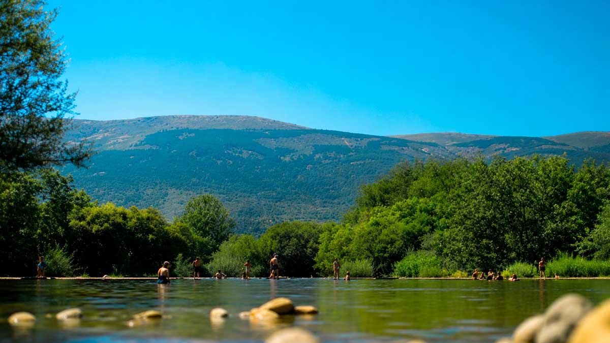 Personas disfrutando en una zona de baño natural en Madrid rodeada de vegetación y montañas